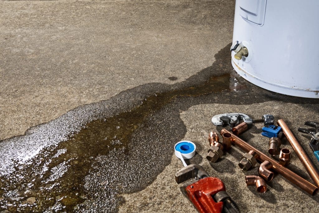 Leaking water heater on concrete floor with plumbing tools and copper pipe fittings nearby.